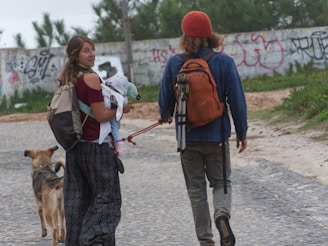 A couple is walking along a cobblestone path near a graffiti-covered wall. The woman is carrying a baby and looking back, while the man carries a large backpack and tripod. A dog is walking beside them.