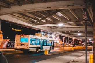 A public bus is driving under an overpass at night, with bright streetlights illuminating the roadway and nearby areas. The background features a dock with a large ship and industrial structures. The environment appears calm and slightly busy, with some traffic signs and barriers visible.