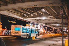 A public bus is driving under an overpass at night, with bright streetlights illuminating the roadway and nearby areas. The background features a dock with a large ship and industrial structures. The environment appears calm and slightly busy, with some traffic signs and barriers visible.