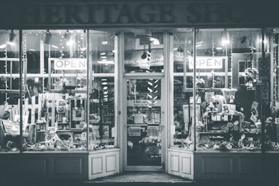 A shopfront with large glass windows showing various items on display. The entrance is adorned with signs indicating 'OPEN' and a variety of goods and decorations can be seen inside, including flowers, wreaths, and other decorative pieces.