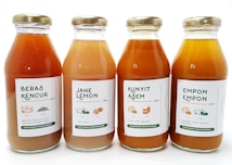 Four glass bottles of traditional Indonesian herbal drinks are aligned side by side. Each bottle has a label with text and small illustrations indicating the drink's ingredients. The labels feature different names: Beras Kencur, Jahe Lemon, Kunyit Asam, and Empon Empon. The bottle caps are metallic and gold in color, and the bottles contain liquids varying from light orange to darker amber.