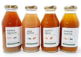 Four glass bottles of traditional Indonesian herbal drinks are aligned side by side. Each bottle has a label with text and small illustrations indicating the drink's ingredients. The labels feature different names: Beras Kencur, Jahe Lemon, Kunyit Asam, and Empon Empon. The bottle caps are metallic and gold in color, and the bottles contain liquids varying from light orange to darker amber.