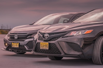 Two black sedans from Honda and Toyota are parked side by side, featuring New Jersey license plates. The lighting is subdued, suggesting a dusk or dawn setting, with soft reflections on the car surfaces.