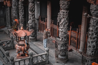 A man stands in a temple-like structure surrounded by intricately carved stone pillars and ornate fencing. The setting includes an incense burner with a decorative roof and a pot placed inside. The floor is made of grayish stone tiles, and the environment has an ancient, historical feel.
