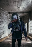 A person wearing a clown mask with curly green hair and a colorful red nose standing in a dimly lit, narrow corridor with concrete walls and ceiling. The figure is dressed in a dark jacket and shirt, with visible blood stains on the hand.