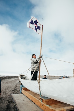 A person is adjusting the sail of a wooden boat placed on a trailer. The boat appears to be on a sandy beach area under a partly cloudy sky. Two other individuals are visible in the background, wearing casual clothing.