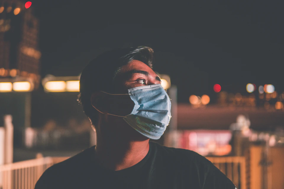 The image shows a man wearing a blue surgical mask, standing on a rooftop at night with buildings and lights visible in the background. The man's gaze is directed upwards towards the sky, suggesting he may be contemplating something or simply enjoying the view.