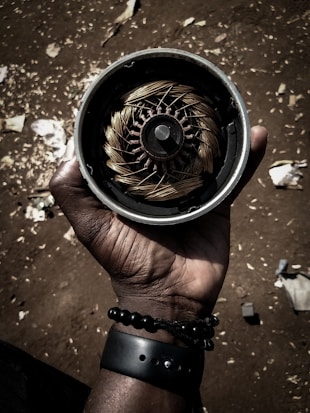A hand is holding a round mechanical device with copper coils and a central metal component, resembling an electric motor or rotor. The ground is visible in the background, scattered with small debris and patches of dirt. The lighting casts strong shadows, giving the image a dramatic effect.