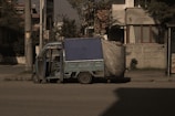 A small, blue motorized rickshaw is parked on an urban street next to a sidewalk. The vehicle has a canopy covering the top and sides, partially made of a tarp material. The surrounding area includes a residential building with patterned walls and windows, as well as utility poles and sparse vegetation.