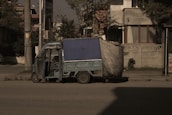 A small, blue motorized rickshaw is parked on an urban street next to a sidewalk. The vehicle has a canopy covering the top and sides, partially made of a tarp material. The surrounding area includes a residential building with patterned walls and windows, as well as utility poles and sparse vegetation.