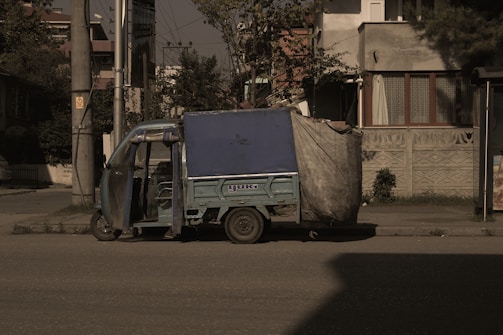 A small, blue motorized rickshaw is parked on an urban street next to a sidewalk. The vehicle has a canopy covering the top and sides, partially made of a tarp material. The surrounding area includes a residential building with patterned walls and windows, as well as utility poles and sparse vegetation.