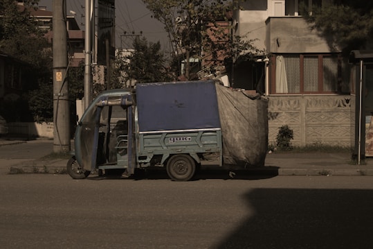A small, blue motorized rickshaw is parked on an urban street next to a sidewalk. The vehicle has a canopy covering the top and sides, partially made of a tarp material. The surrounding area includes a residential building with patterned walls and windows, as well as utility poles and sparse vegetation.