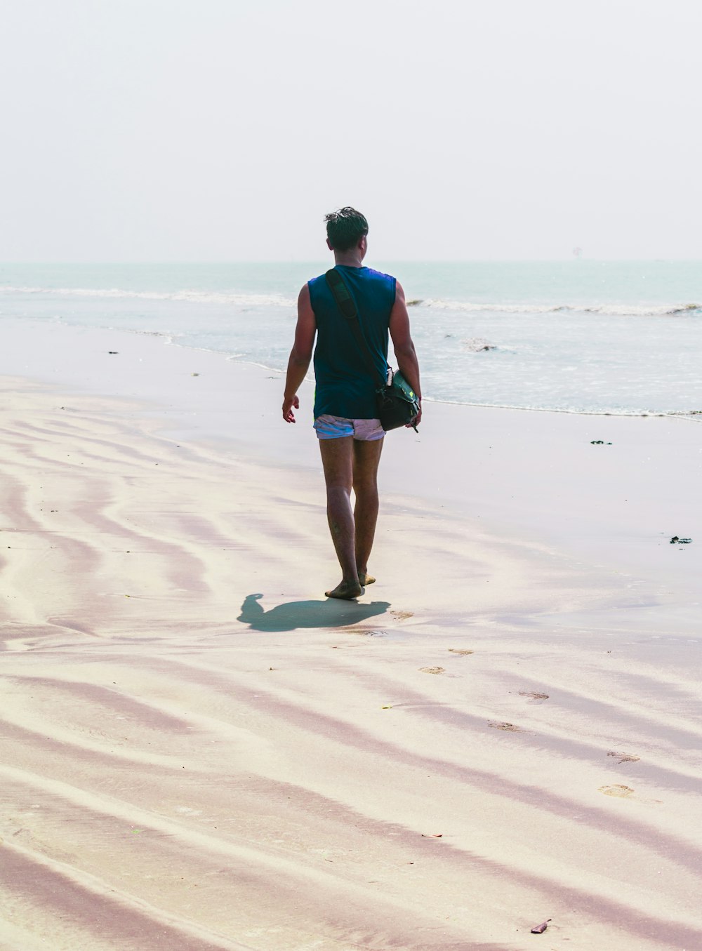 파란색 셔츠와 검은 반바지를 입은 남자가 낮 동안 해변을 걷고 있다