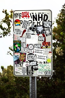 A metal sign has been covered with a variety of colorful and monochrome stickers featuring text, logos, and graphics. The stickers are layered and overlap each other, showcasing different designs and artistic styles. The background shows some greenery and tree foliage.