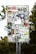 A metal sign has been covered with a variety of colorful and monochrome stickers featuring text, logos, and graphics. The stickers are layered and overlap each other, showcasing different designs and artistic styles. The background shows some greenery and tree foliage.