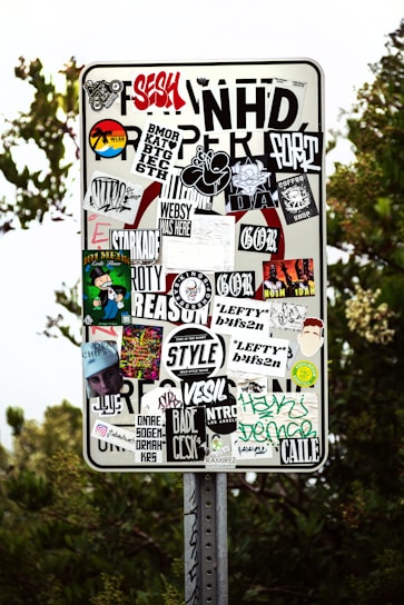 A metal sign has been covered with a variety of colorful and monochrome stickers featuring text, logos, and graphics. The stickers are layered and overlap each other, showcasing different designs and artistic styles. The background shows some greenery and tree foliage.