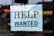 A sign with the words 'Help Wanted' is taped to a glass window. The sign is made of light blue paper with bold black lettering, and it appears to be indicating a job advertisement or hiring notice.