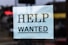 A sign with the words 'Help Wanted' is taped to a glass window. The sign is made of light blue paper with bold black lettering, and it appears to be indicating a job advertisement or hiring notice.
