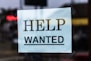 A sign with the words 'Help Wanted' is taped to a glass window. The sign is made of light blue paper with bold black lettering, and it appears to be indicating a job advertisement or hiring notice.