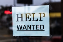 A sign with the words 'Help Wanted' is taped to a glass window. The sign is made of light blue paper with bold black lettering, and it appears to be indicating a job advertisement or hiring notice.