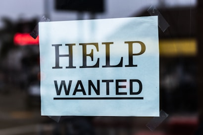 A sign with the words 'Help Wanted' is taped to a glass window. The sign is made of light blue paper with bold black lettering, and it appears to be indicating a job advertisement or hiring notice.
