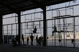 Silhouetted workers are constructing a large building with scaffolding in the foreground. Various people are positioned at different levels on the scaffolding, wearing helmets and safety gear. The setting appears industrial or commercial, with a clear sky in the background and a ship visible at the dock.