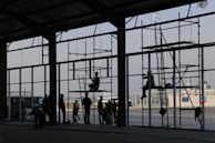 Silhouetted workers are constructing a large building with scaffolding in the foreground. Various people are positioned at different levels on the scaffolding, wearing helmets and safety gear. The setting appears industrial or commercial, with a clear sky in the background and a ship visible at the dock.