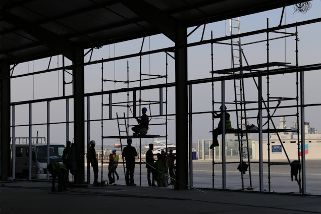 Silhouetted workers are constructing a large building with scaffolding in the foreground. Various people are positioned at different levels on the scaffolding, wearing helmets and safety gear. The setting appears industrial or commercial, with a clear sky in the background and a ship visible at the dock.