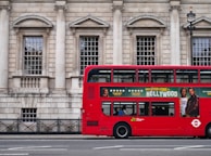 A red double-decker bus is parked alongside a historic stone building with large windows and columns. The side of the bus features an advertisement for a movie with star ratings and images of actors. A few people are visible inside the bus through the windows.