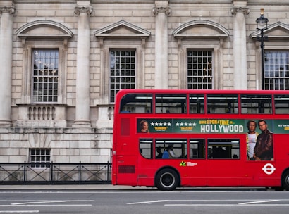 A red double-decker bus is parked alongside a historic stone building with large windows and columns. The side of the bus features an advertisement for a movie with star ratings and images of actors. A few people are visible inside the bus through the windows.