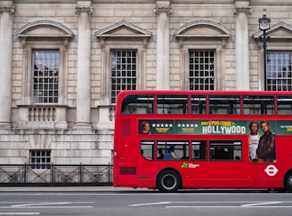 A red double-decker bus is parked alongside a historic stone building with large windows and columns. The side of the bus features an advertisement for a movie with star ratings and images of actors. A few people are visible inside the bus through the windows.