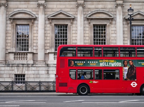 A red double-decker bus is parked alongside a historic stone building with large windows and columns. The side of the bus features an advertisement for a movie with star ratings and images of actors. A few people are visible inside the bus through the windows.