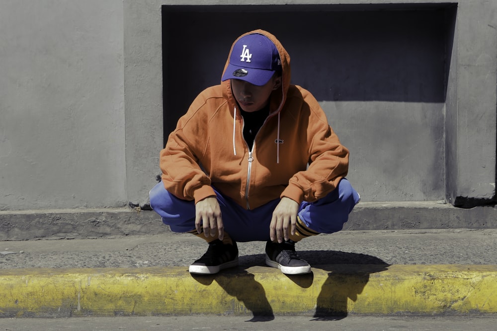 boy in orange zip up hoodie and black pants sitting on gray concrete floor