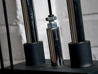 Close-up of a metallic mechanical component with cylindrical rods and nuts, set against a textured white brick wall.