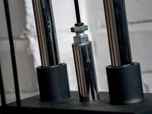 Close-up of a metallic mechanical component with cylindrical rods and nuts, set against a textured white brick wall.