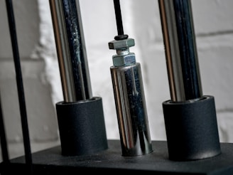 Close-up of a metallic mechanical component with cylindrical rods and nuts, set against a textured white brick wall.