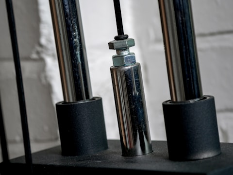 Close-up of a metallic mechanical component with cylindrical rods and nuts, set against a textured white brick wall.