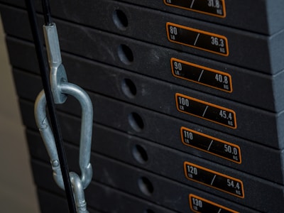 A stack of gym weight plates marked with both pounds and kilograms, connected by a metal chain or hook. The numbers range from 80 to 130 pounds or 36.3 to 54.4 kilograms.