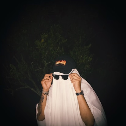 A person is draped with a white sheet, resembling a ghost, while wearing sunglasses and a black cap with a logo. The background is dark, with faint outlines of foliage visible.