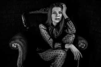 A woman with long hair is seated on an ornate armchair with one hand resting on her head. She is wearing a sheer polka-dot blouse and appears contemplative. The setting is dark, enhancing the contrast between her light clothing and the background.