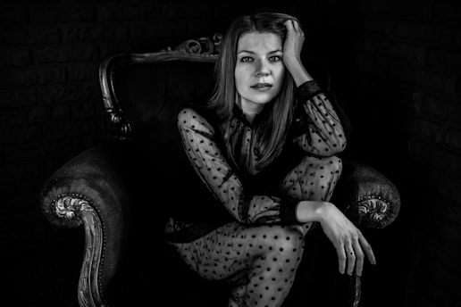 A woman with long hair is seated on an ornate armchair with one hand resting on her head. She is wearing a sheer polka-dot blouse and appears contemplative. The setting is dark, enhancing the contrast between her light clothing and the background.