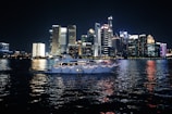A luxurious yacht sails on a calm body of water against the backdrop of a vibrant city skyline illuminated at night. The high-rise buildings are adorned with bright neon lights, reflecting vivid colors on the water surface.