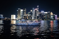 A luxurious yacht sails on a calm body of water against the backdrop of a vibrant city skyline illuminated at night. The high-rise buildings are adorned with bright neon lights, reflecting vivid colors on the water surface.