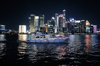 A luxurious yacht sails on a calm body of water against the backdrop of a vibrant city skyline illuminated at night. The high-rise buildings are adorned with bright neon lights, reflecting vivid colors on the water surface.