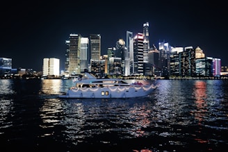 A luxurious yacht sails on a calm body of water against the backdrop of a vibrant city skyline illuminated at night. The high-rise buildings are adorned with bright neon lights, reflecting vivid colors on the water surface.