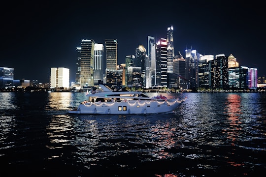 A luxurious yacht sails on a calm body of water against the backdrop of a vibrant city skyline illuminated at night. The high-rise buildings are adorned with bright neon lights, reflecting vivid colors on the water surface.