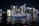 A luxurious yacht sails on a calm body of water against the backdrop of a vibrant city skyline illuminated at night. The high-rise buildings are adorned with bright neon lights, reflecting vivid colors on the water surface.