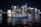 A luxurious yacht sails on a calm body of water against the backdrop of a vibrant city skyline illuminated at night. The high-rise buildings are adorned with bright neon lights, reflecting vivid colors on the water surface.