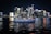 A luxurious yacht sails on a calm body of water against the backdrop of a vibrant city skyline illuminated at night. The high-rise buildings are adorned with bright neon lights, reflecting vivid colors on the water surface.