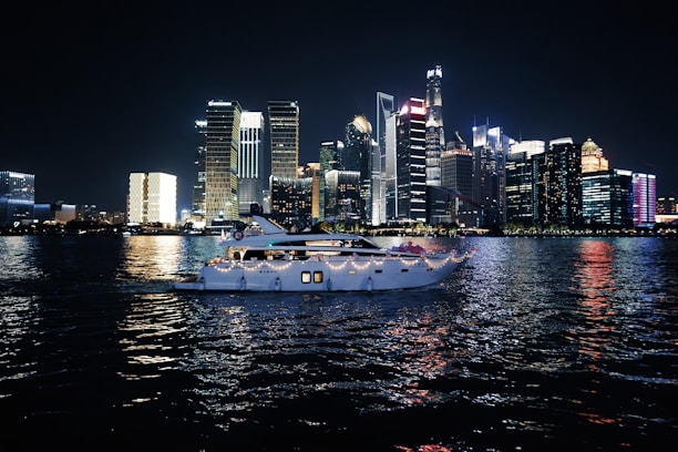 A luxurious yacht sails on a calm body of water against the backdrop of a vibrant city skyline illuminated at night. The high-rise buildings are adorned with bright neon lights, reflecting vivid colors on the water surface.